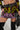 A person wearing a brightly colored, patterned "Too Hot To Handle Embellished Mini Skirt" paired with a matching top and a studded belt. They hold a rhinestone-studded black handbag and are wearing black knee-high boots. The outfit features abstract designs in pink, green, and black.