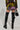 A person with long hair is wearing a vibrant sequined outfit that includes a TOO HOT TO HANDLE Embellished Mini Skirt, paired with black knee-high boots decorated with chains and buckles. They are holding a small faux leather handbag featuring metal studs, posing against a plain background.
