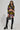 A person with long braids is wearing futuristic sunglasses, a colorful patterned faux leather crop top, and a TOO HOT TO HANDLE EMBELLISHED MINI SKIRT, paired with studded thigh-high black boots. They are holding a rhinestone-studded handbag and stand confidently in front of a plain background.