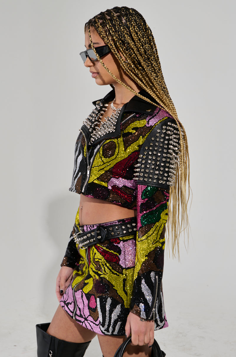 A person with long braided hair wearing sunglasses and a Too Hot to Handle Embellished Moto adorned with metallic studs. The colorful, patterned outfit features a faux leather collar, a cropped jacket, and a matching skirt paired with knee-high black boots against a plain white background.