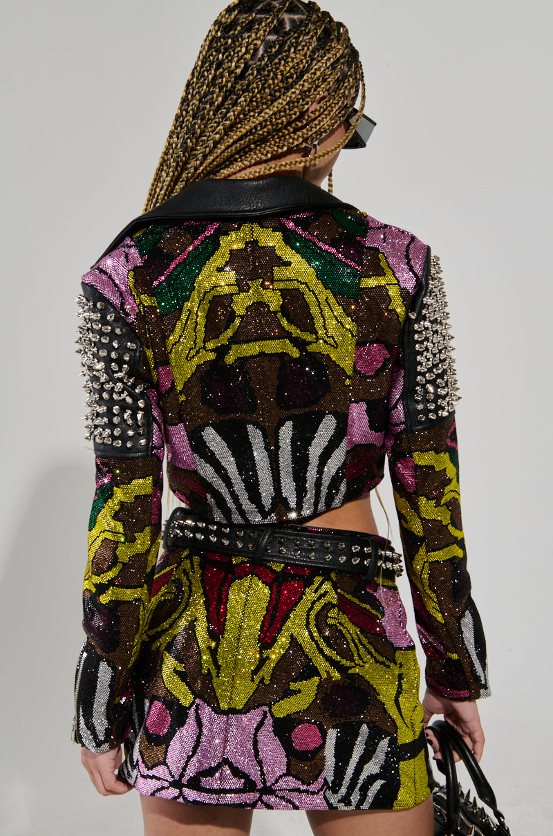 A person with braided hair and sunglasses faces away, showing off a vibrant, sequined outfit in pink, green, and gold with studded shoulder details. The ensemble mirrors the Too Hot to Handle Embellished Moto design as they hold a matching bag, completing their bold fashion statement.
