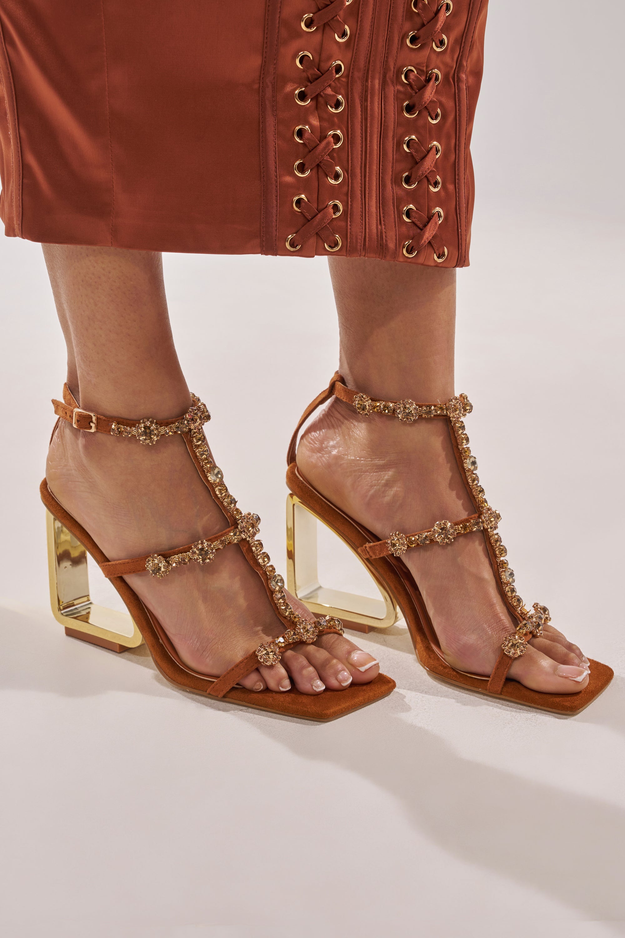 Wearing rust-colored pants with gold grommets, a person models the AZALEA WANG SCHANTELLE Tan Embellished Upper Wedge Sandal, which features a comfort cushion insole and beaded strap details, against a light background. 0