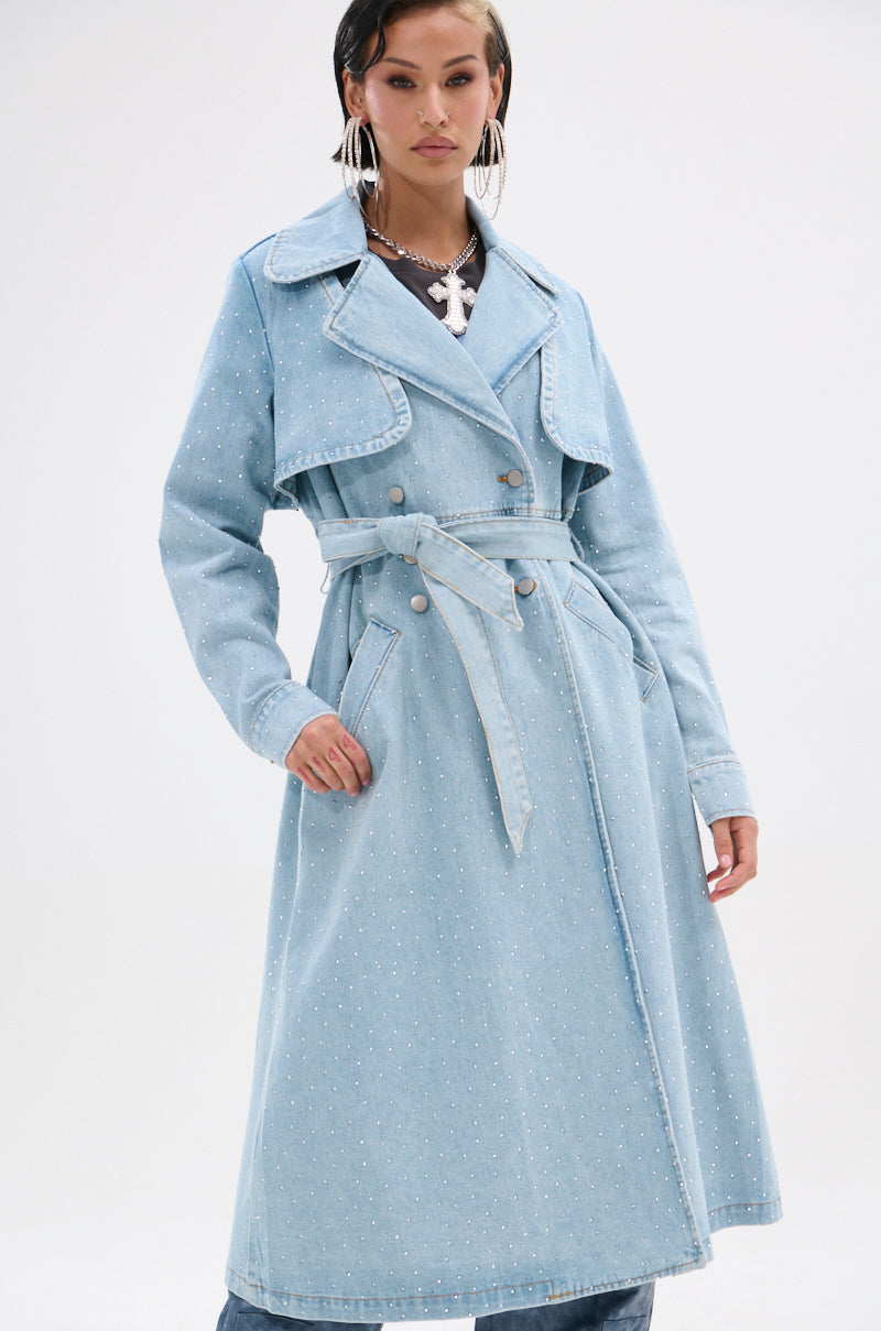 A person wearing the REYA RHINESTONE DENIM TRENCH—a light blue, vintage-wash, oversized double-breasted coat with subtle embellishments—stands against a plain white background.