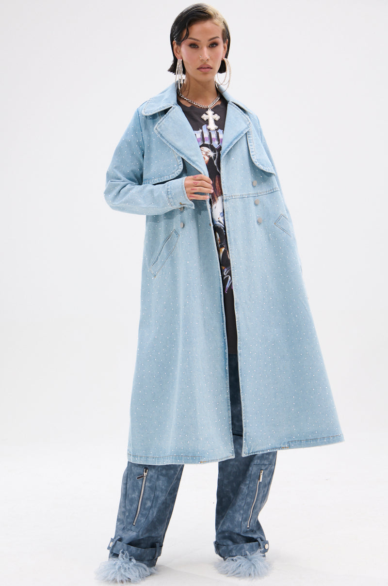 A woman stands confidently in the REYA RHINESTONE DENIM TRENCH, a vintage-wash long denim coat with small studs, wide lapels, and flap pockets, styled with blue zippered pants, furry shoes, slicked-back hair, and bold earrings.
