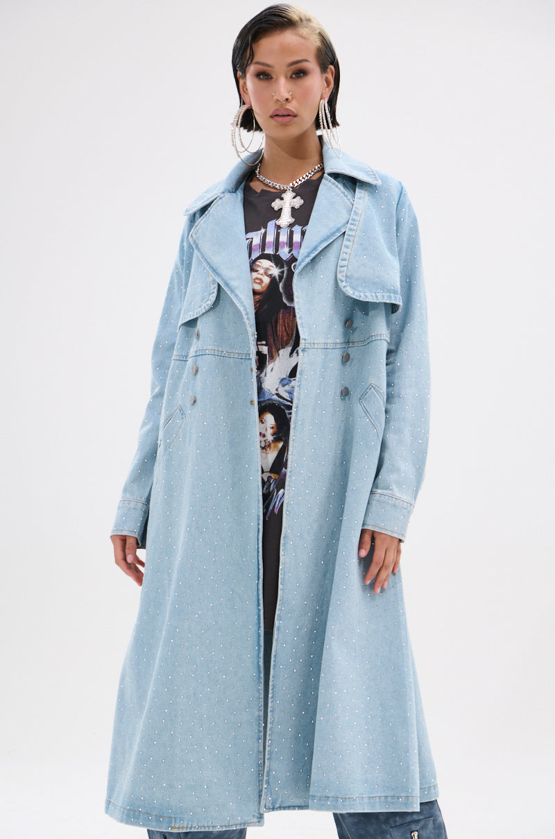 Wearing the REYA RHINESTONE DENIM TRENCH in light blue with silver embellishments over a graphic tee, accessorized with large hoop earrings and layered necklaces, the person stands against a plain white background.