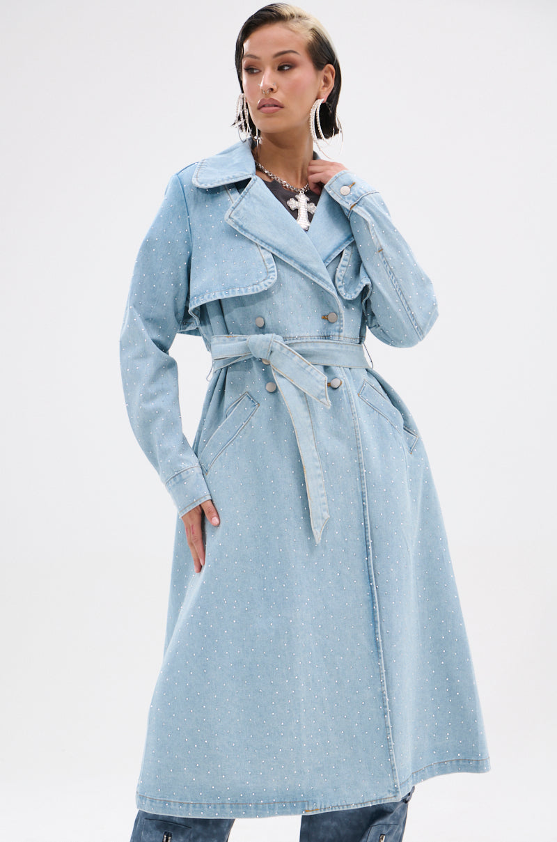 A woman models the REYA RHINESTONE DENIM TRENCH, a long, vintage-wash trench coat with a belt and subtle rhinestone details, against a plain white background.