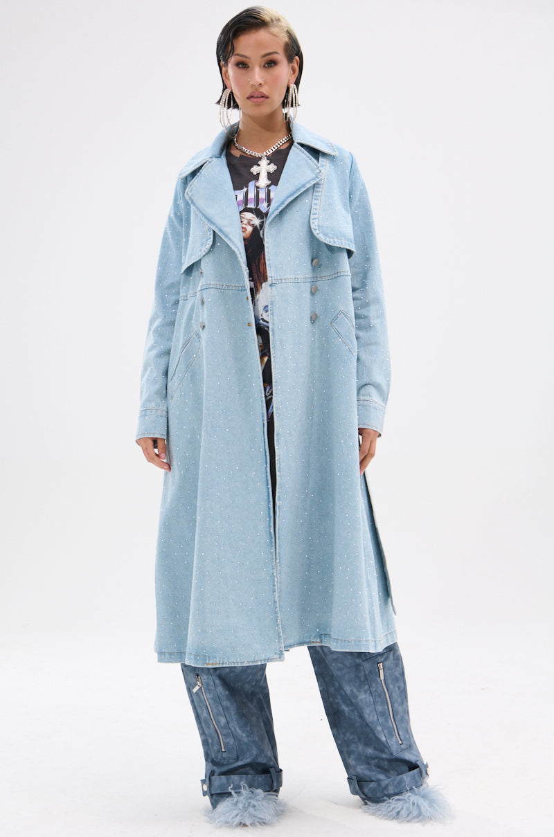 A woman wears the REYA RHINESTONE DENIM TRENCH—an oversized, light blue rhinestone denim coat—over a graphic shirt and blue textured pants with zippered, fur-trimmed cuffs. She stands facing forward against a plain white background.