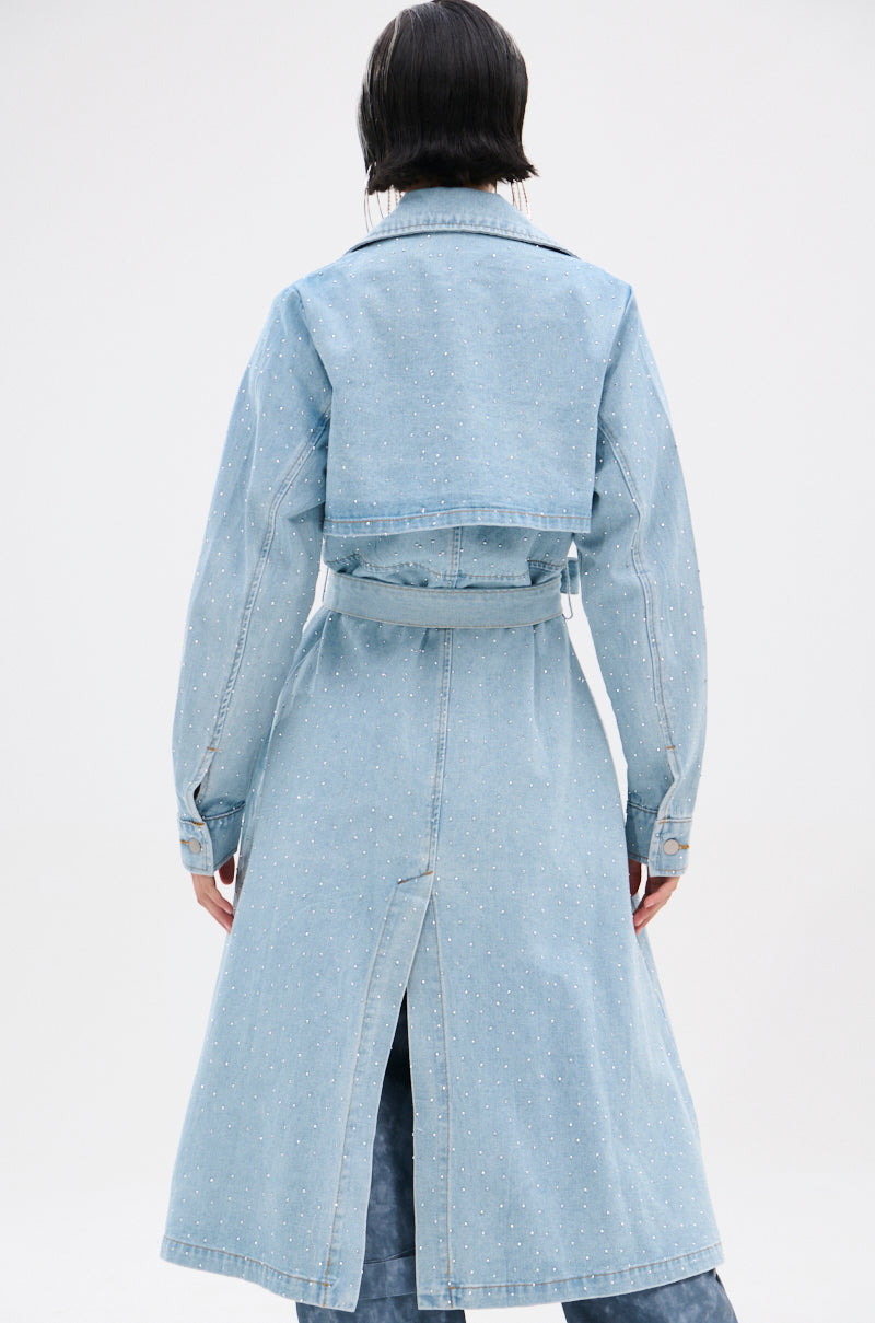 A person with short dark hair wears the REYA RHINESTONE DENIM TRENCH—an oversized, vintage-wash denim coat with small rhinestones, a belt, and back slit—shown from the back against a plain white background.