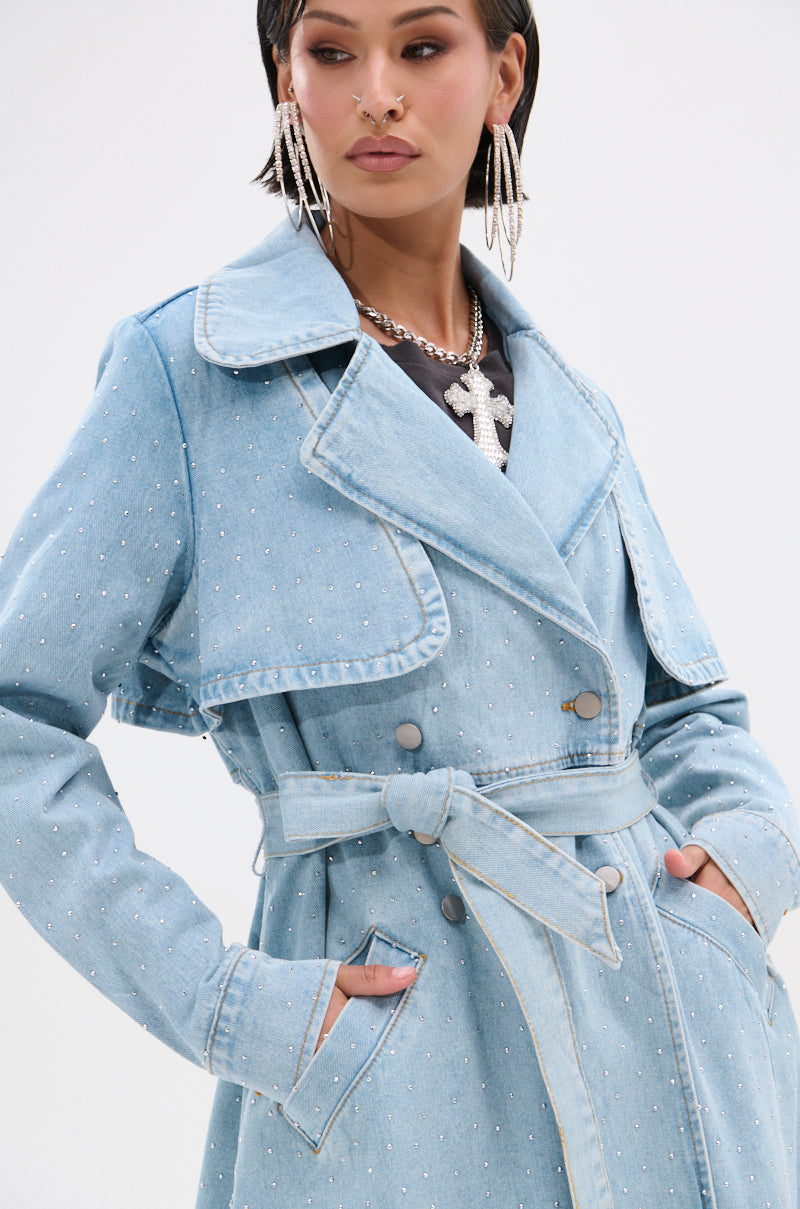 A woman models the REYA RHINESTONE DENIM TRENCH, an oversized light blue vintage-wash denim coat with silver studs and a tie belt. She has short dark hair, statement earrings, a cross necklace, and her hands in the pockets.