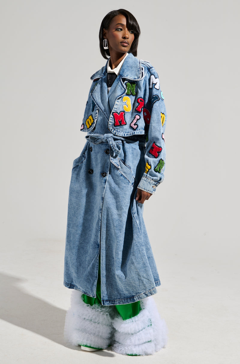 A person wears the TAKE NOTES DENIM TRENCH embellished with vibrant alphabet patches, complemented by oversized green pants and fluffy white boots against a plain, light-colored background.
