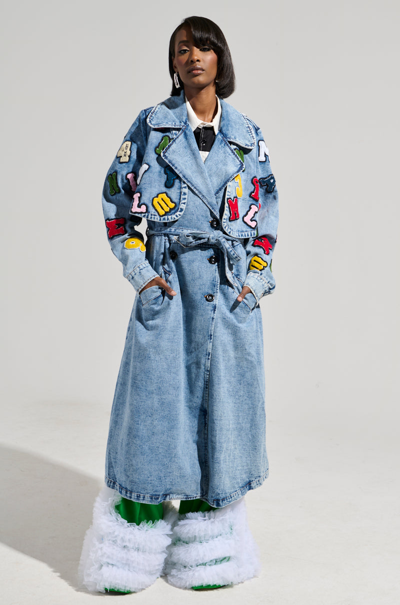 A person stands confidently in the TAKE NOTES DENIM TRENCH, featuring alphabet patches. They pair it with white furry boots and green pants, set against a plain background that highlights the maxi-length silhouette and makes a bold fashion statement.