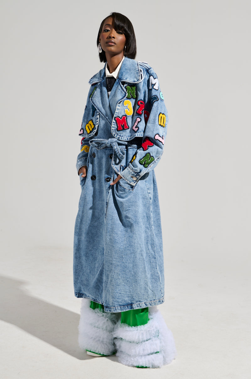 A person confidently wears the TAKE NOTES DENIM TRENCH, adorned with colorful alphabet patches. They pair it with bright green pants and fluffy white boots, creating a striking look against the plain light gray background.
