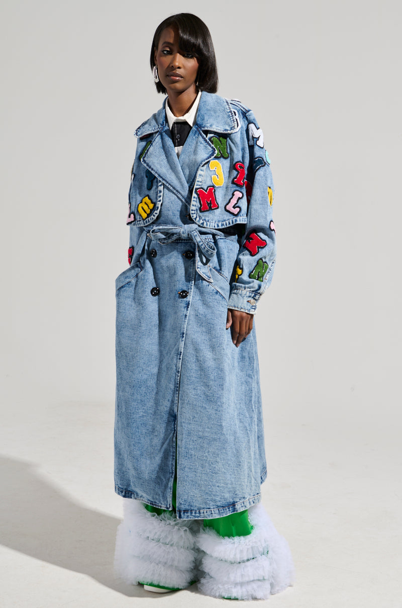 Dressed in the TAKE NOTES DENIM TRENCH, which features a maxi-length silhouette with eye-catching alphabet patches and a tie belt, they style it with bright green leggings and fluffy white boots. The setting is plain and neutral.