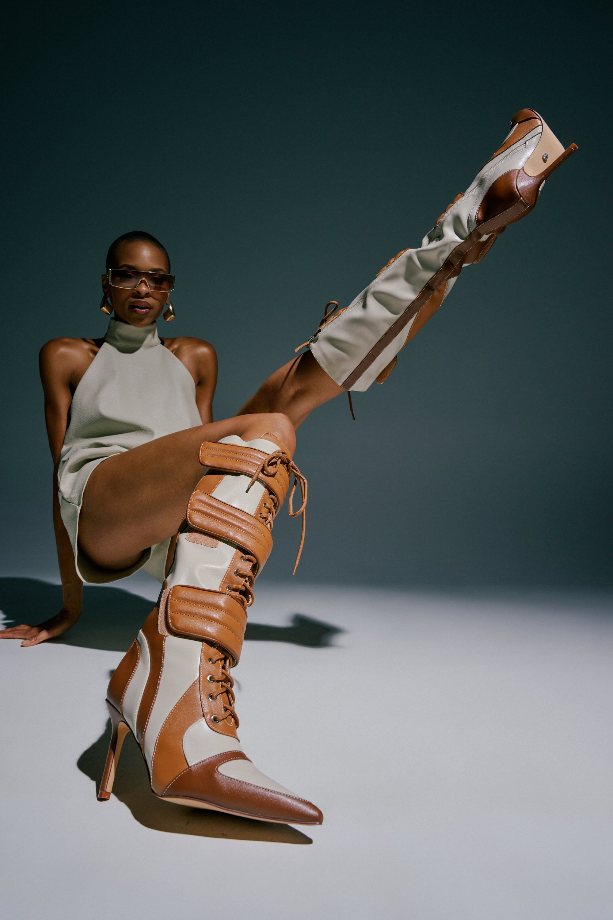 A person poses confidently, wearing a sleeveless outfit and bold AZALEA WANG SCHUMACHER TAN STILETTO BOOTS—knee-high with a striking faux leather paneled design in tan and white. With one leg raised high, the statement footwear stands out against the neutral background.