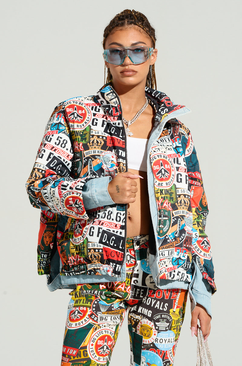 A woman with braided hair wears a RED LIGHT LAYERED GRAPHIC BOMBER and matching pants adorned with bold prints and logos. The oversized fit jacket is paired with a white crop top, and she carries a small bag. Her look stands out against a plain, light-colored background.