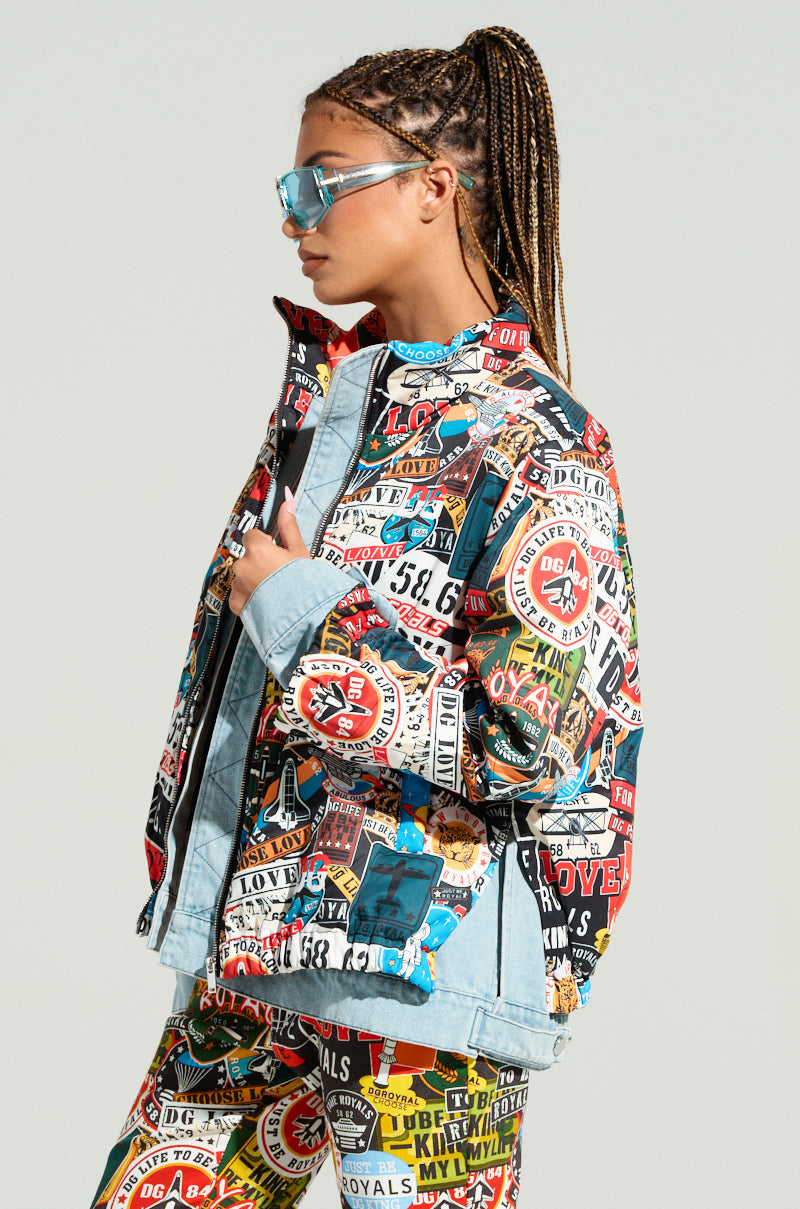 A person with braided hair, wearing reflective sunglasses, poses in a colorful RED LIGHT LAYERED GRAPHIC BOMBER and matching pants adorned with various logos and text. The jacket features an oversized fit and a multi-layered design. The background is light gray.