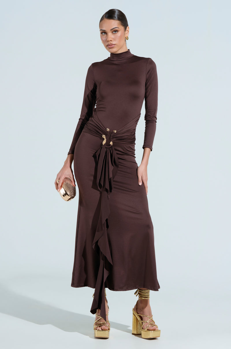 A woman confidently wears THE BEST ONE YET brown maxi dress with a high neckline and ruffle details. She carries a gold metallic snake clutch, wears gold platform heels, and accessorizes with sleekly styled hair and gold earrings.