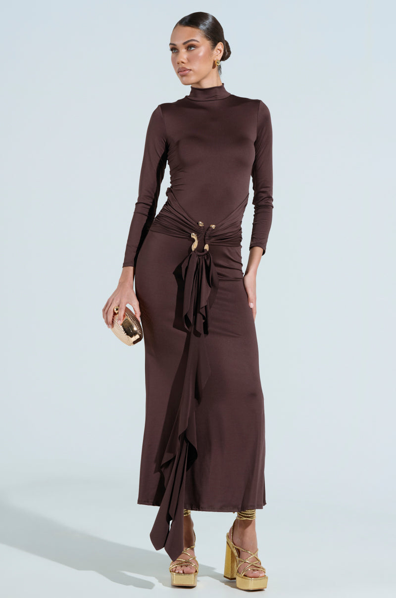 A woman models "THE BEST ONE YET" brown maxi dress featuring a high neckline and asymmetric draping, made from stretchy mesh. She accessorizes with a small gold clutch and gold platform sandals, her hair styled in a sleek bun.