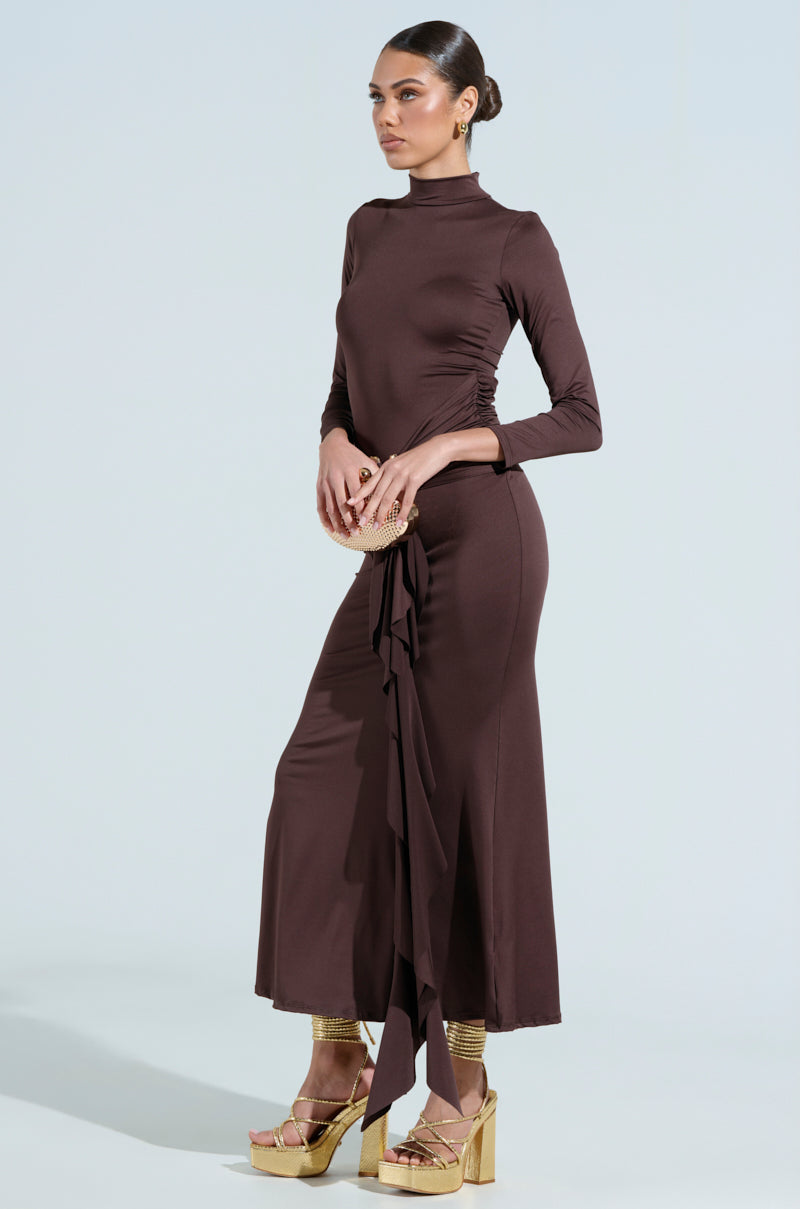 A woman confidently poses in "The Best One Yet" brown maxi dress, crafted from stretchy mesh. She holds a beige clutch and wears gold snake platform heels, with her hair in a bun, set against a light blue backdrop.