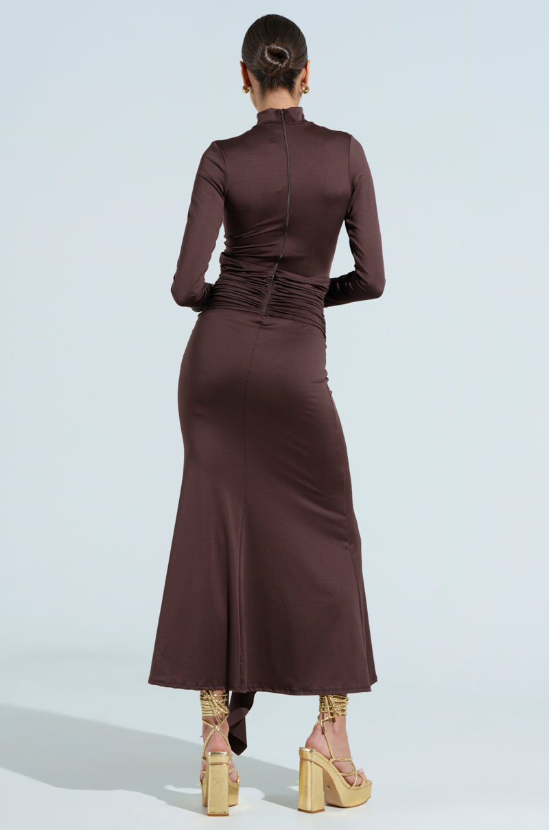 In THE BEST ONE YET BROWN MAXI DRESS, a person stands with their back to the camera. Styled in an updo, they wear platform heels. The long-sleeved, stretchy brown mesh dress gracefully flares at the bottom like a gold metallic snake as it extends to their ankles.