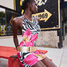 A woman in the WILD THANG PRINTED MINI DRESS with a bold zebra and pink floral print plus a wide gold belt sits on a red picnic bench. Behind her, a mural reads “WE BUILT Gold! SINCE 1969” with an arrow pointing right.