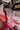 A woman with short hair and bold makeup poses outdoors on a red picnic table, wearing the WILD THANG PRINTED MINI DRESS—featuring a tropical zebra print with pink flowers, layered necklaces, a wide studded belt, and a mural wall in the background.