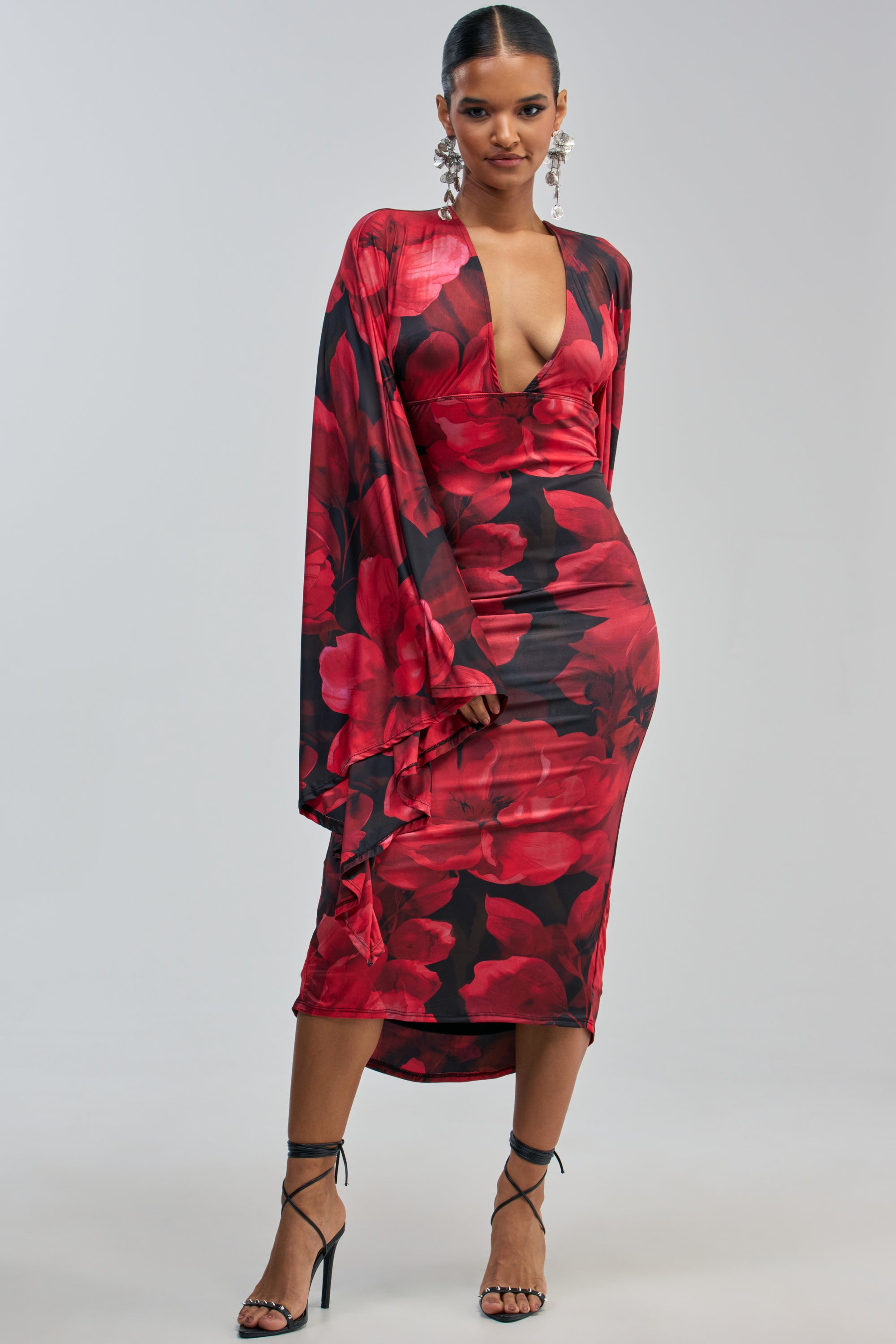 Wearing the SHINY LIKE ME MAXI DRESS IN RED FLORAL—a red floral-print maxi dress featuring a deep V-neck and flared sleeves—paired with black strappy heels, this woman stands confidently against a plain light gray background, showcasing an elegant evening look.