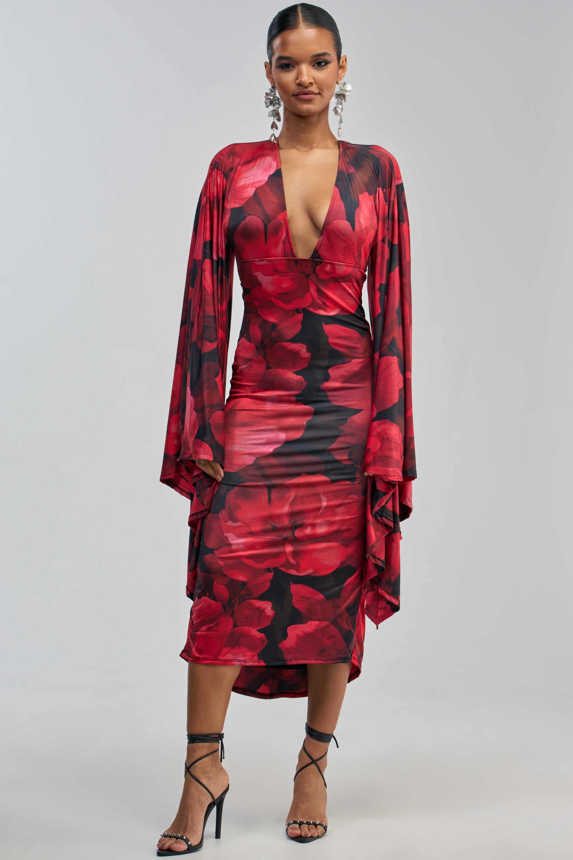 A woman showcases the SHINY LIKE ME MAXI DRESS IN RED FLORAL, featuring long, flowing sleeves and a deep V-neckline. She pairs the stunning red floral evening dress with strappy black heels and statement earrings, standing against a plain light background—making it the perfect choice for any formal occasion.