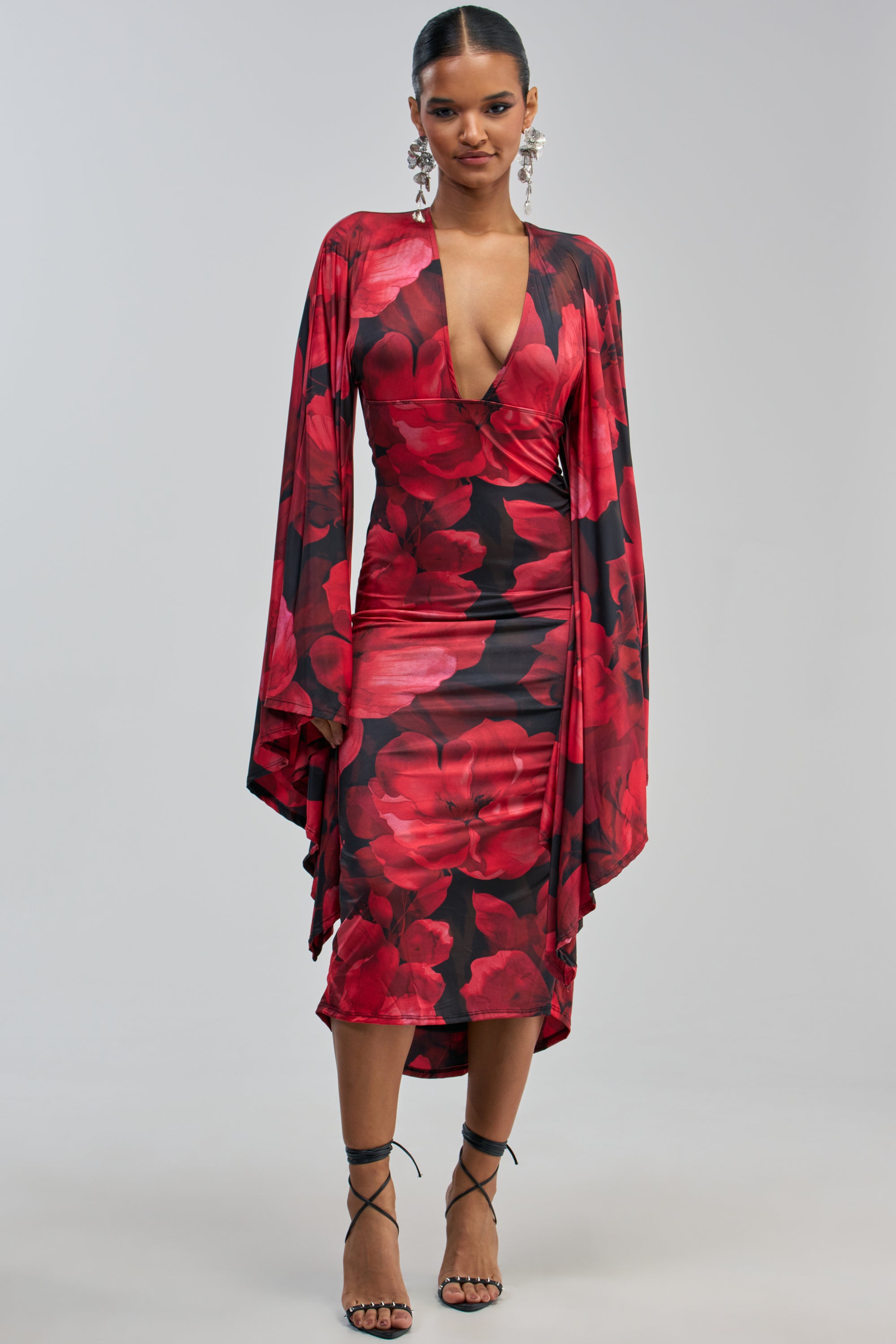 A woman wears the SHINY LIKE ME MAXI DRESS IN RED FLORAL, featuring long, draped sleeves and a deep V neckline, paired with black strappy heels, standing against a plain light gray background.