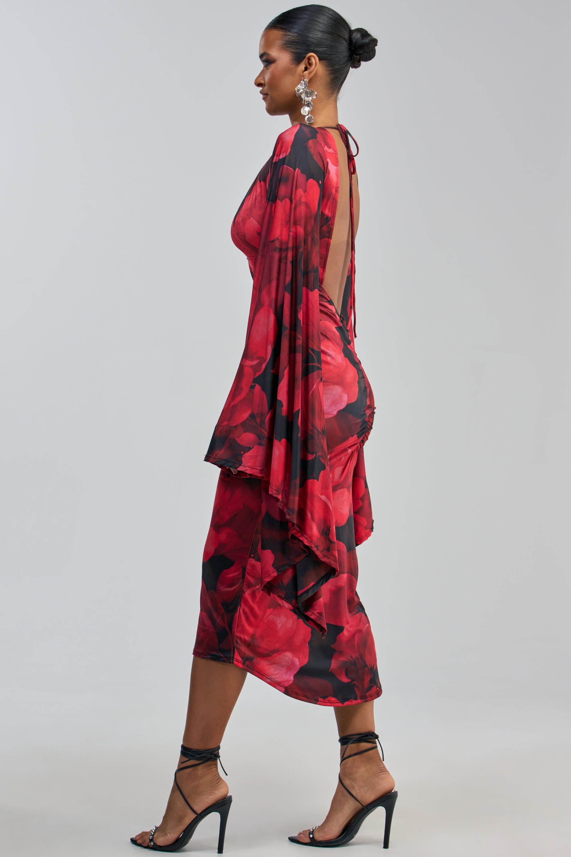 Wearing the SHINY LIKE ME MAXI DRESS IN RED FLORAL, a woman stands in profile, showcasing its open back and draped fabric. She completes her look with high-heeled sandals and statement earrings.
