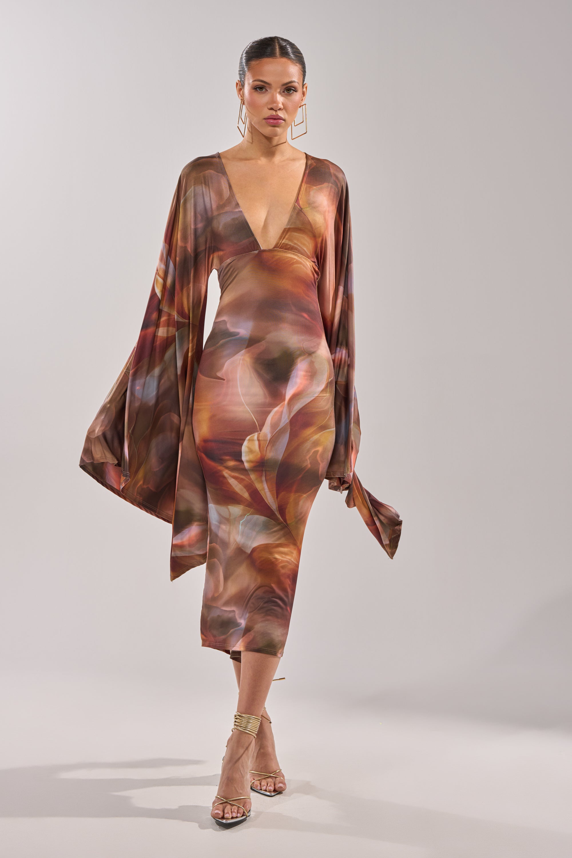 Wearing the SHINY LIKE ME MAXI DRESS with wide sleeves and a plunge neckline, a woman stands against a light background, paired with gold geometric earrings and strappy heels. 2