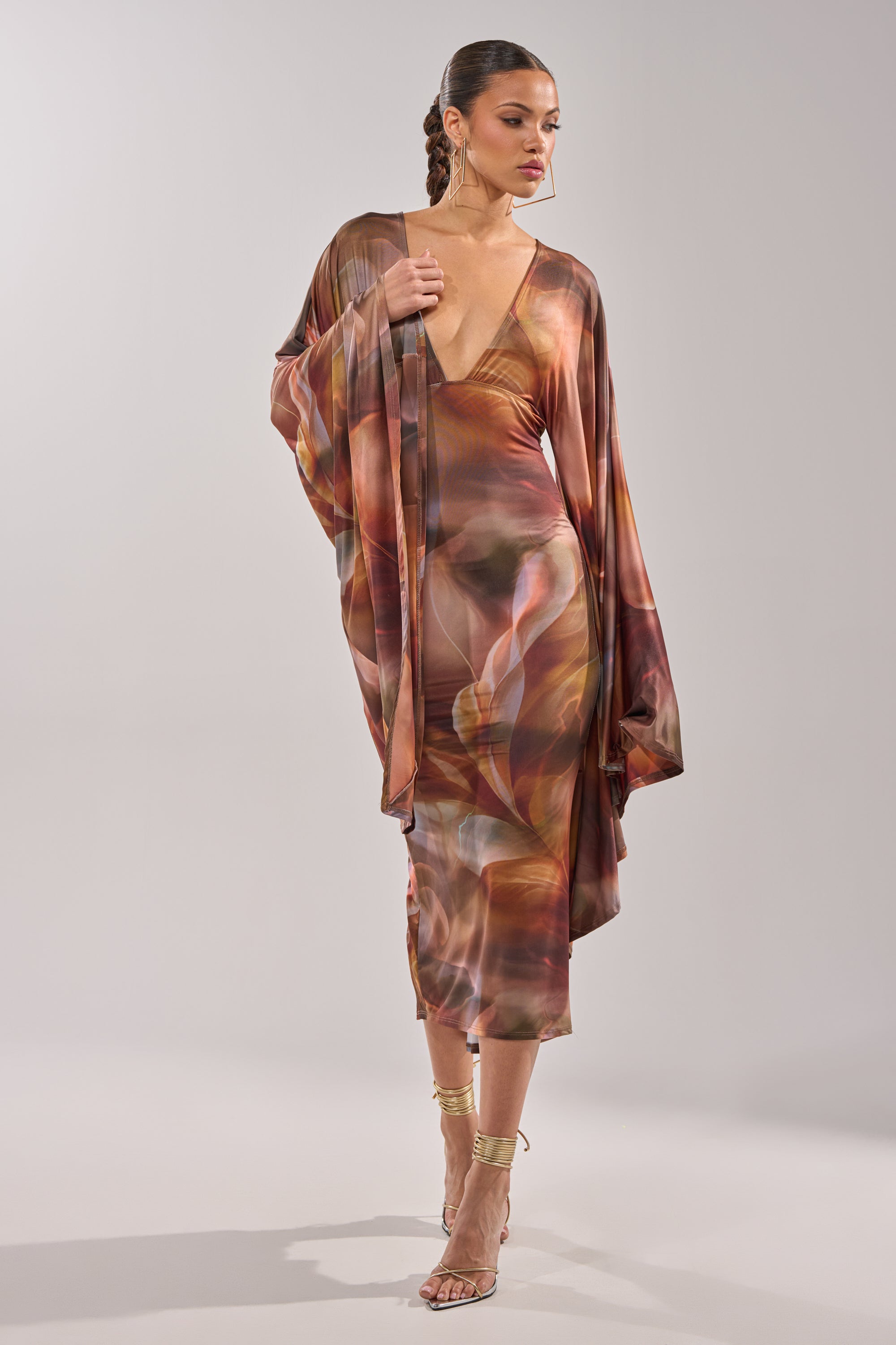 A woman wears the SHINY LIKE ME MAXI DRESS, featuring a flowing, abstract print, long sleeves, a deep V-neckline, and a body-skimming silhouette. She pairs it with strappy heels and styled braids against a plain light background. 3