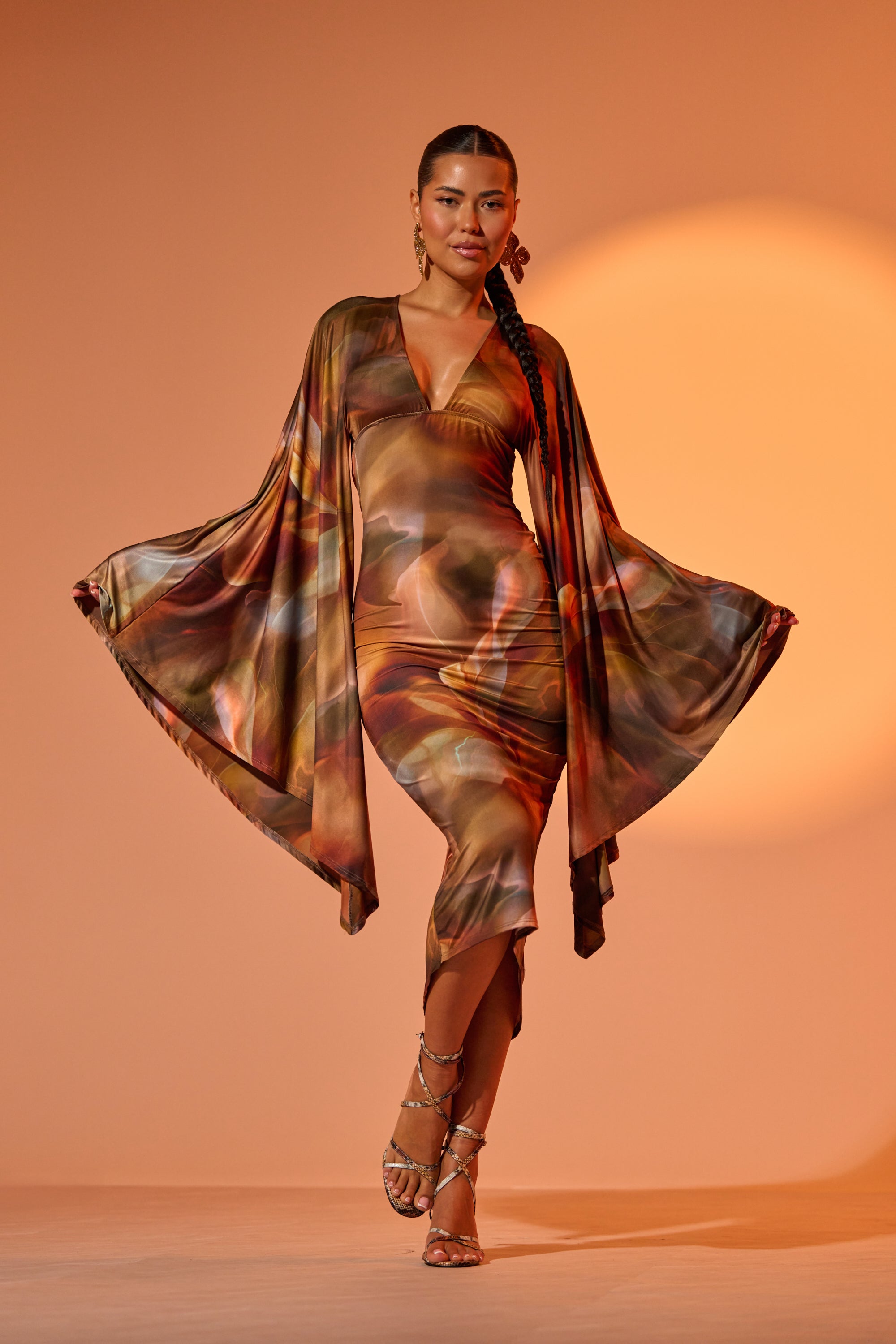 A woman poses confidently in the SHINY LIKE ME MAXI DRESS, featuring long, wing-like sleeves and a deep plunge neckline, paired with strappy heels. Her hair is styled in a long braid against an orange background. 1