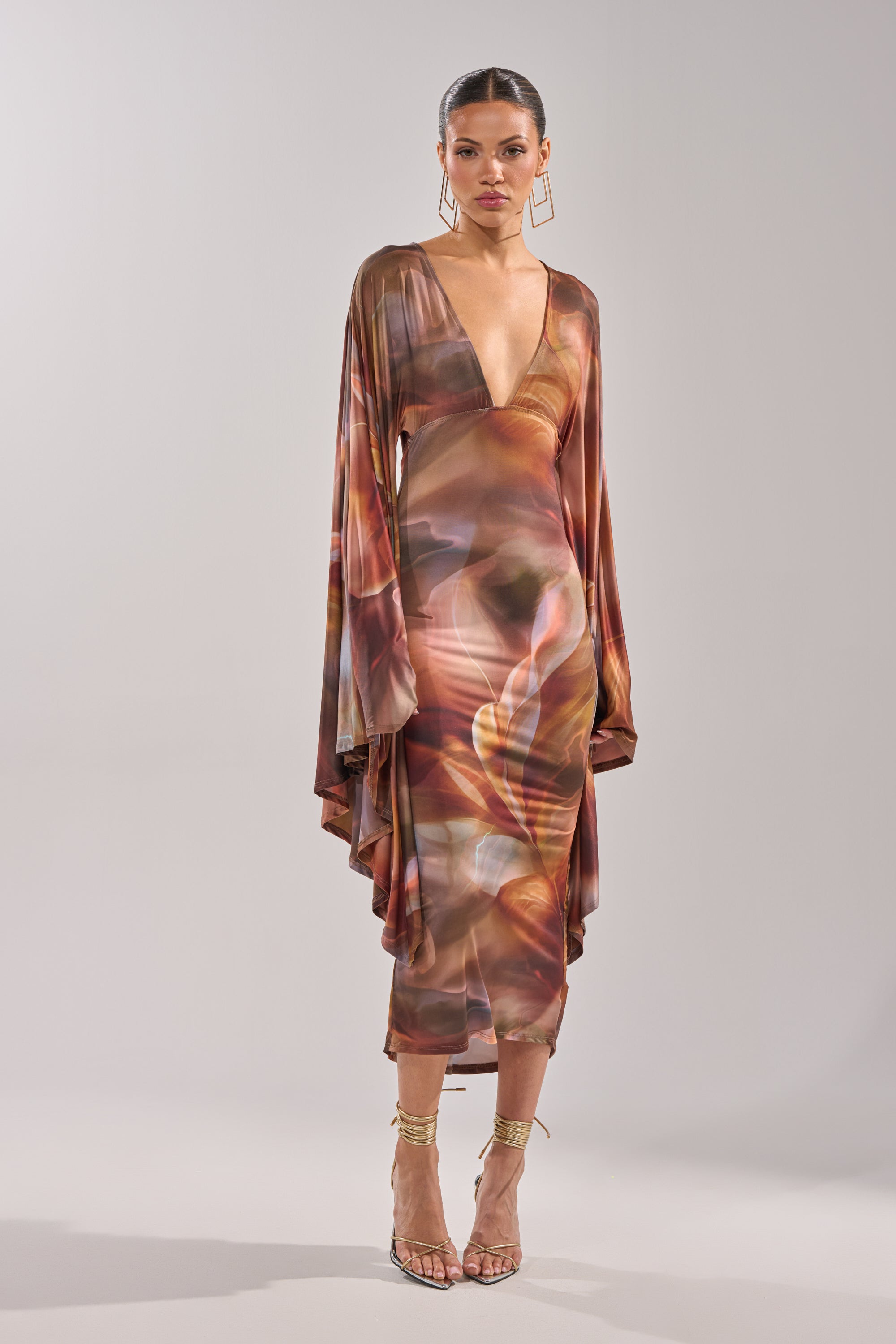 A woman poses against a plain light background wearing the SHINY LIKE ME MAXI DRESS, a long, flowy maxi dress with an abstract print, deep V-neck, wide sleeves, and gold strappy high-heeled sandals. 4