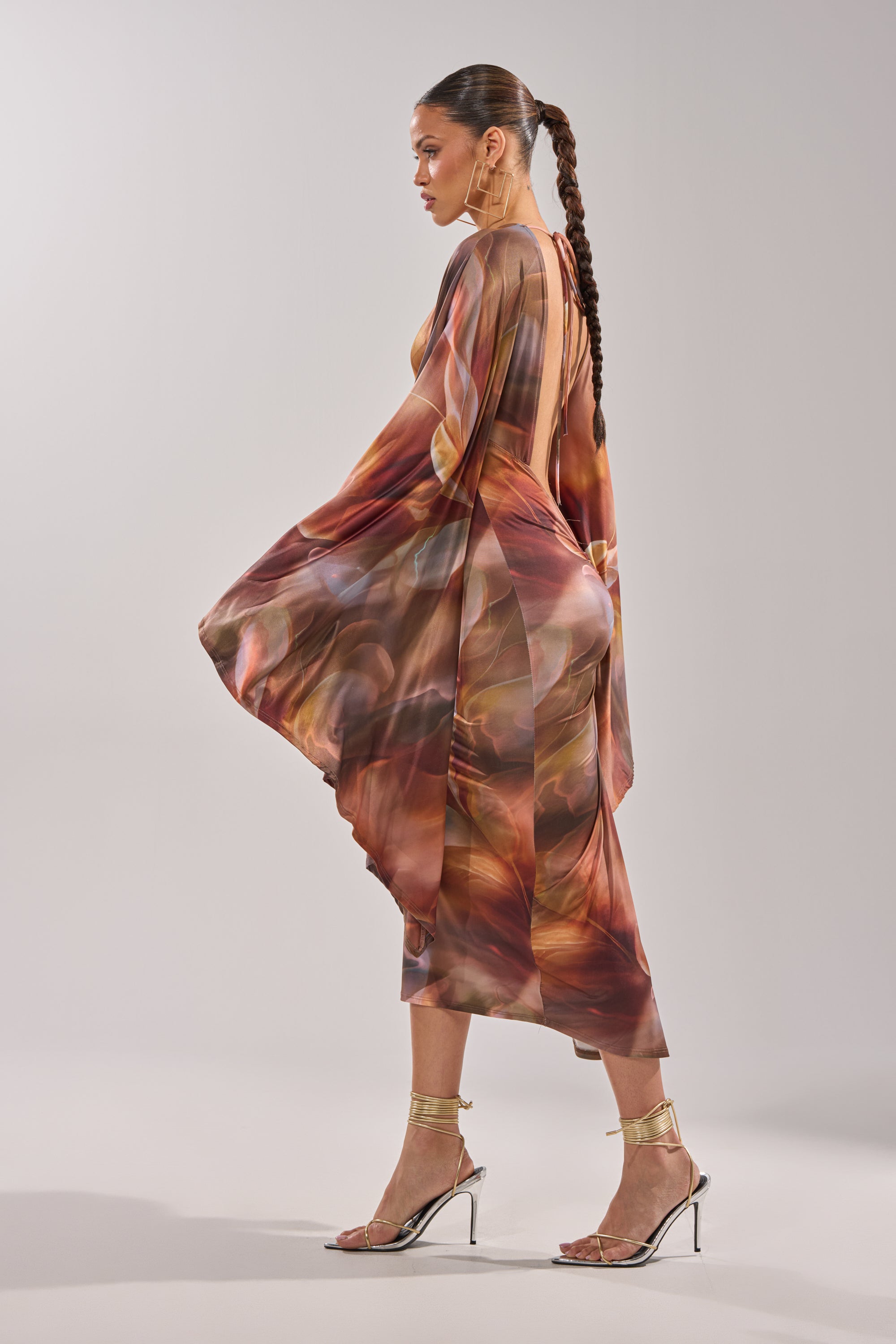 A woman with a long braided ponytail stands sideways against a plain light background, wearing the SHINY LIKE ME MAXI DRESS with an abstract print, open back, and gold strappy high-heeled sandals. 5