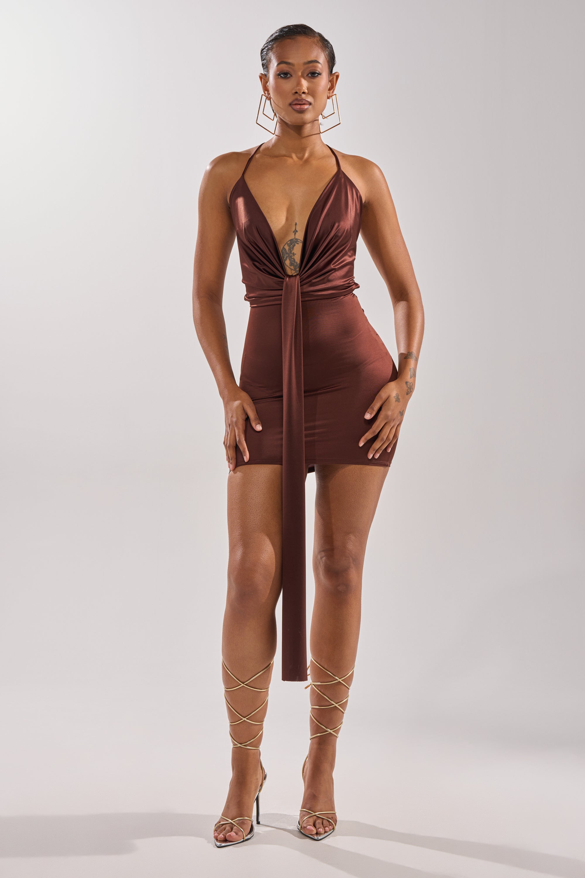 A woman exudes confidence in the BODY TEA MINI DRESS—a fitted deep brown mini with a plunging neckline and front tie. She pairs it with large hoop earrings, strappy heels, and shows off a chest tattoo against a plain light background.