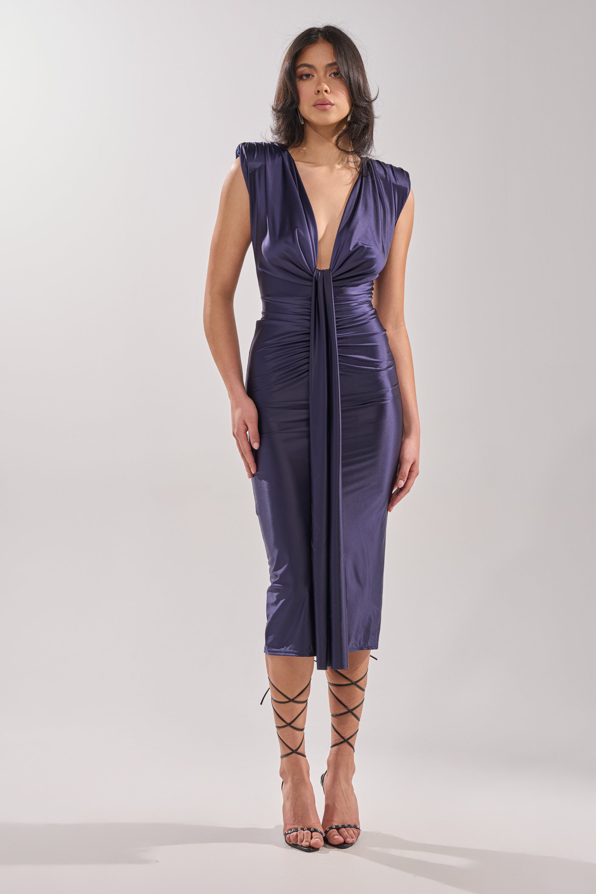 A woman stands against a plain background wearing the FLY ME OUT SATIN MAXI DRESS, a sleeveless deep v-neck purple satin maxi with ruched detailing and knot-front draped tie, styled with strappy black heels. 5