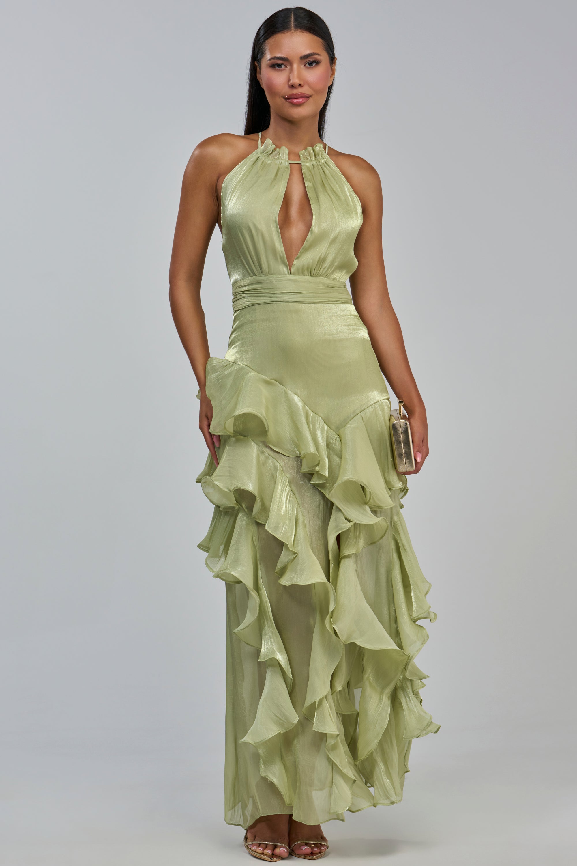 A woman wearing the HONORABLE MENTIONS CHIFFON MAXI DRESS IN SAGE—a light green, halter-neck gown with a plunging neckline and a ruffled, layered skirt—holds a small clutch while standing against a plain light gray background, making it an ideal choice for a wedding guest dress.