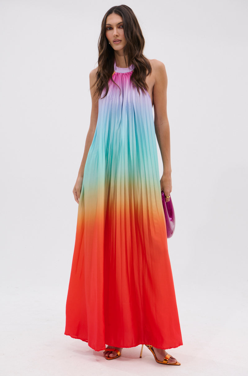 A woman wears the SUNSET PLEATED OMBRE MAXI DRESS, featuring a sleeveless design with rainbow ombre pleated satin. She accessorizes with a purple clutch and heeled sandals.