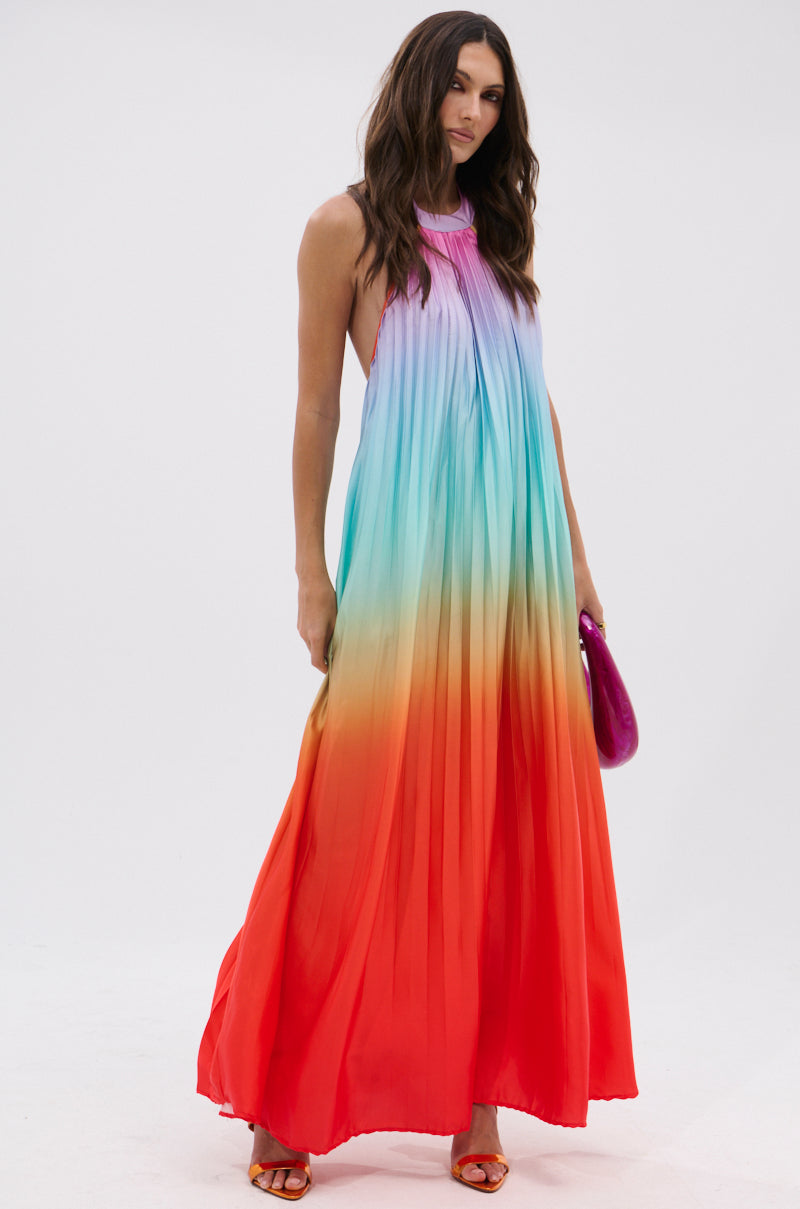 A woman wears the SUNSET PLEATED OMBRE MAXI DRESS, a sleeveless satin maxi with pleats and a purple-to-red rainbow gradient. She accessorizes with a purple clutch bag and orange sandals.