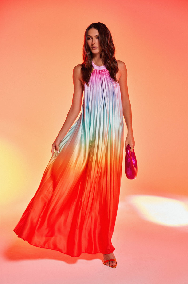 A woman with long, wavy dark hair stands against an orange background wearing the SUNSET PLEATED OMBRE MAXI DRESS with a colorful design, holding a shiny purple clutch.