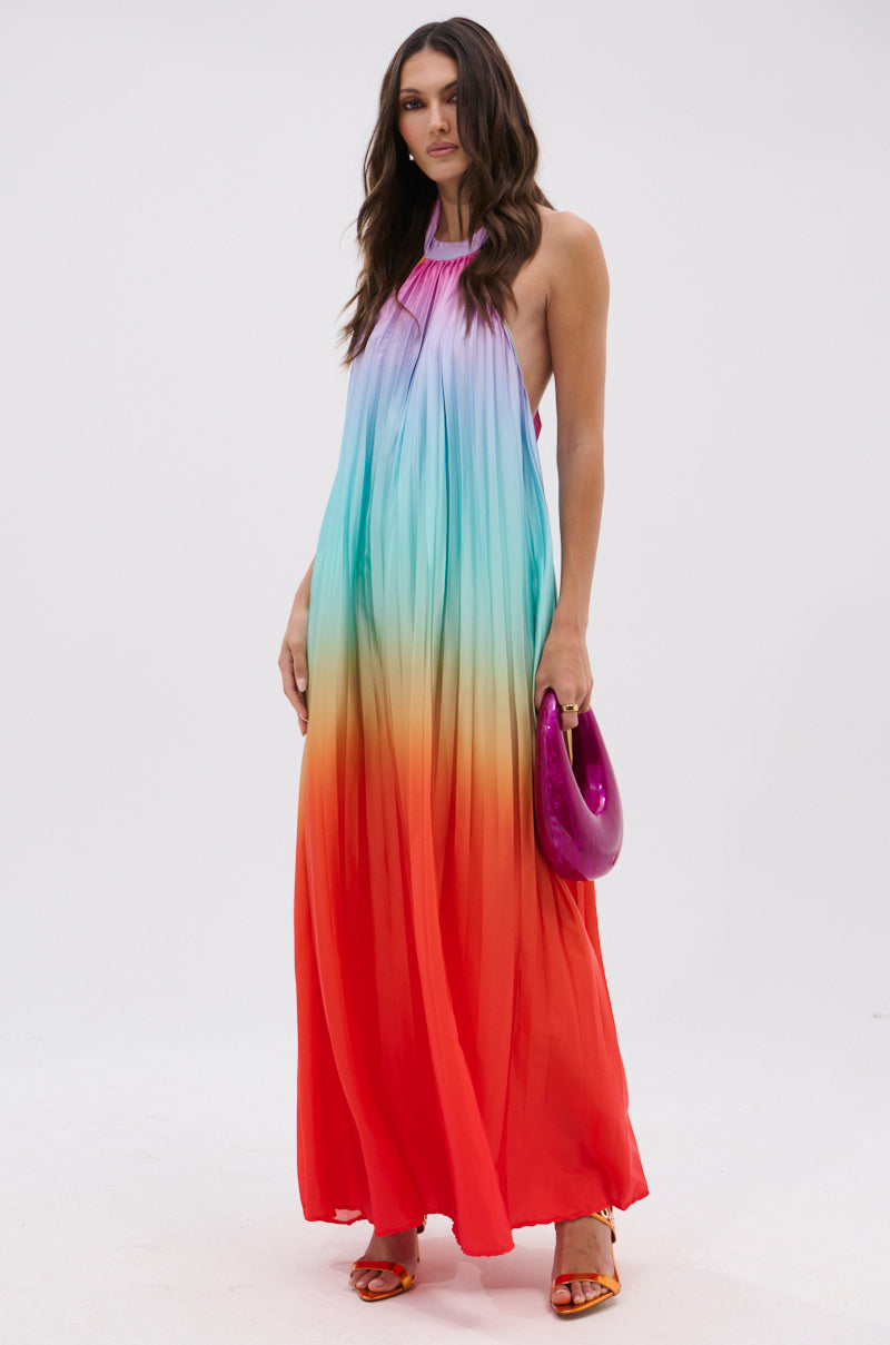 A woman in the SUNSET PLEATED OMBRE MAXI DRESS, featuring a halter neckline and pastel blue-to-red gradient, stands against a white background holding a shiny purple clutch and wearing orange heels.