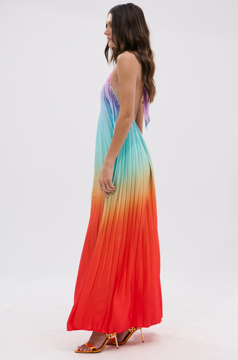 Woman stands sideways in the SUNSET PLEATED OMBRE MAXI DRESS with shades of purple, blue, green, yellow, and red. The dress features an open back and thin straps, paired with orange heeled sandals against a plain background.