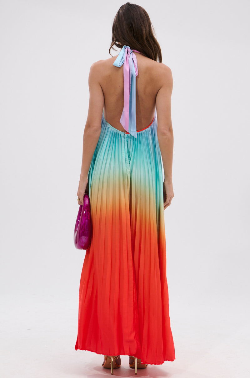 A woman with long, wavy brown hair faces away, wearing the SUNSET PLEATED OMBRE MAXI DRESS featuring a pleated rainbow gradient from blue to red and a halter neckline. She carries a shiny pink clutch.