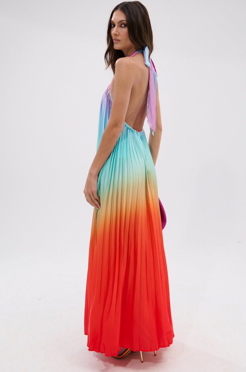 A woman stands sideways wearing the SUNSET PLEATED OMBRE MAXI DRESS, featuring a halter neckline and gradient rainbow pleats that fade from light blue at the top to bright red at the hem, set against a plain white background.