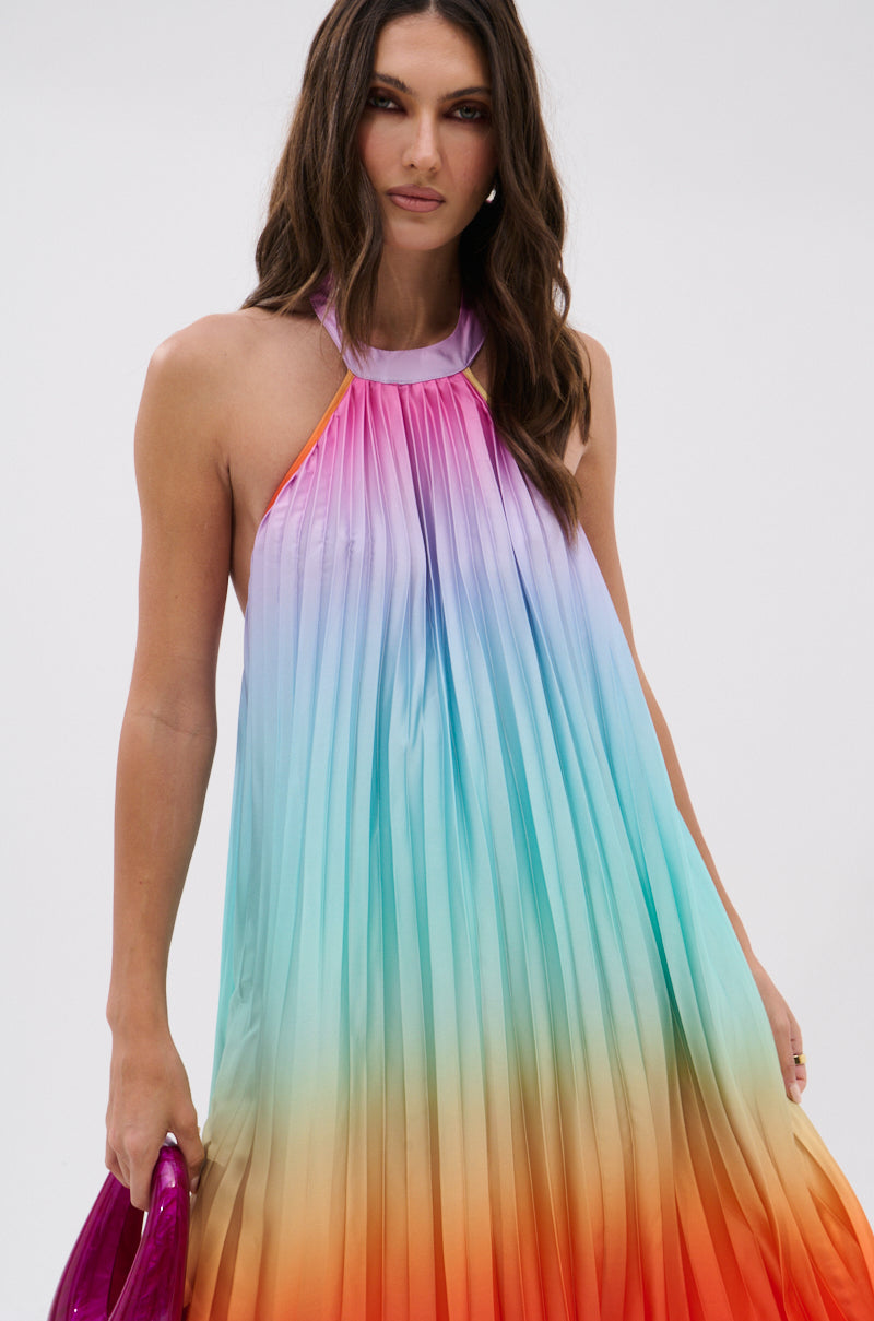 A woman with long brown hair models the SUNSET PLEATED OMBRE MAXI DRESS, featuring a halter neckline and a pleated gradient from purple to red, and holds a shiny pink clutch against a plain background.