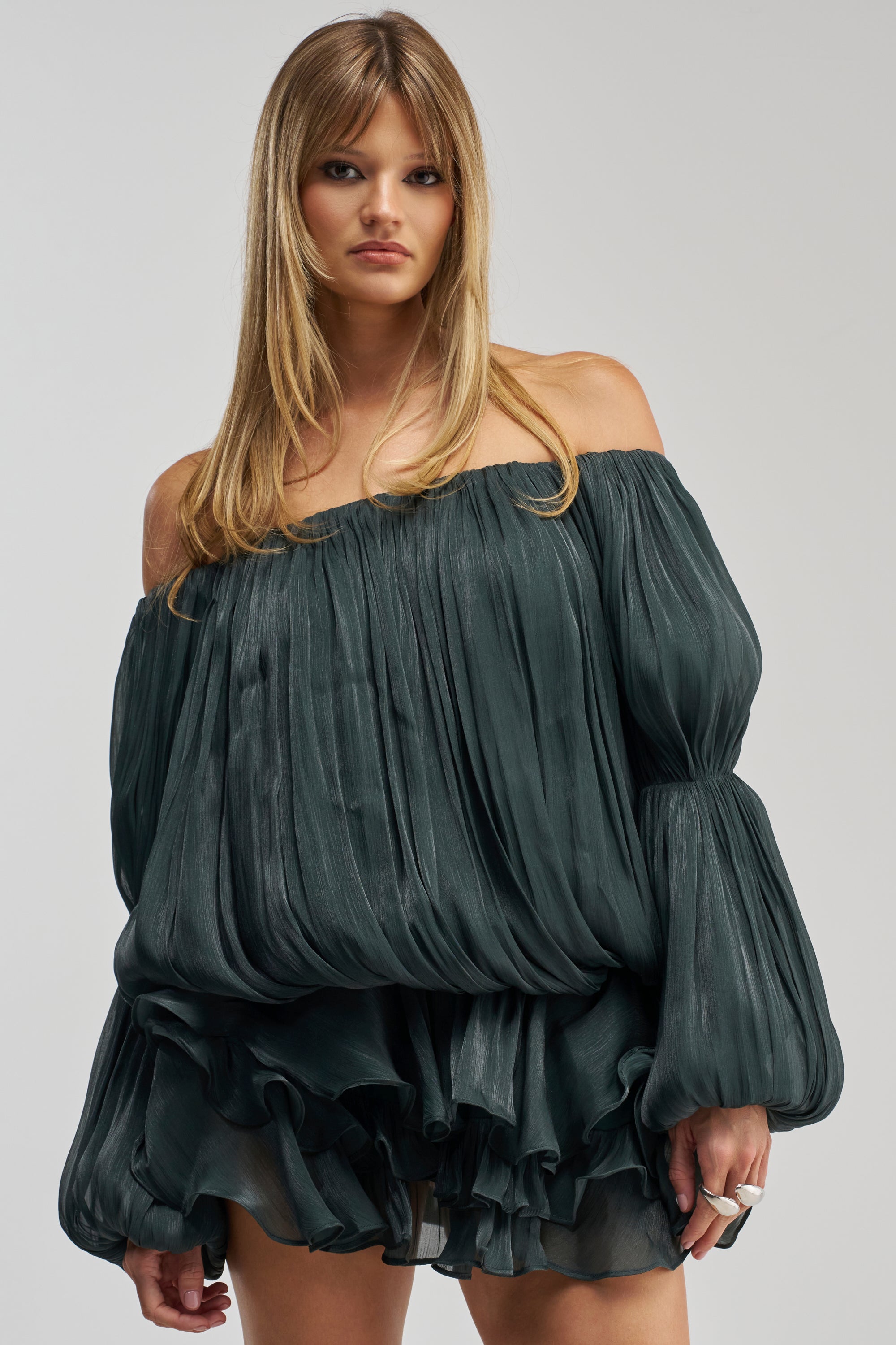 A woman with long blonde hair wears the FAVORITE DAUGHTER Chiffon Mini Dress in Deep Sea, an off-the-shoulder style crafted from dark green chiffon with voluminous sleeves and a ruffled, pleated skirt. She stands against a plain light gray background, looking at the camera with a neutral expression.