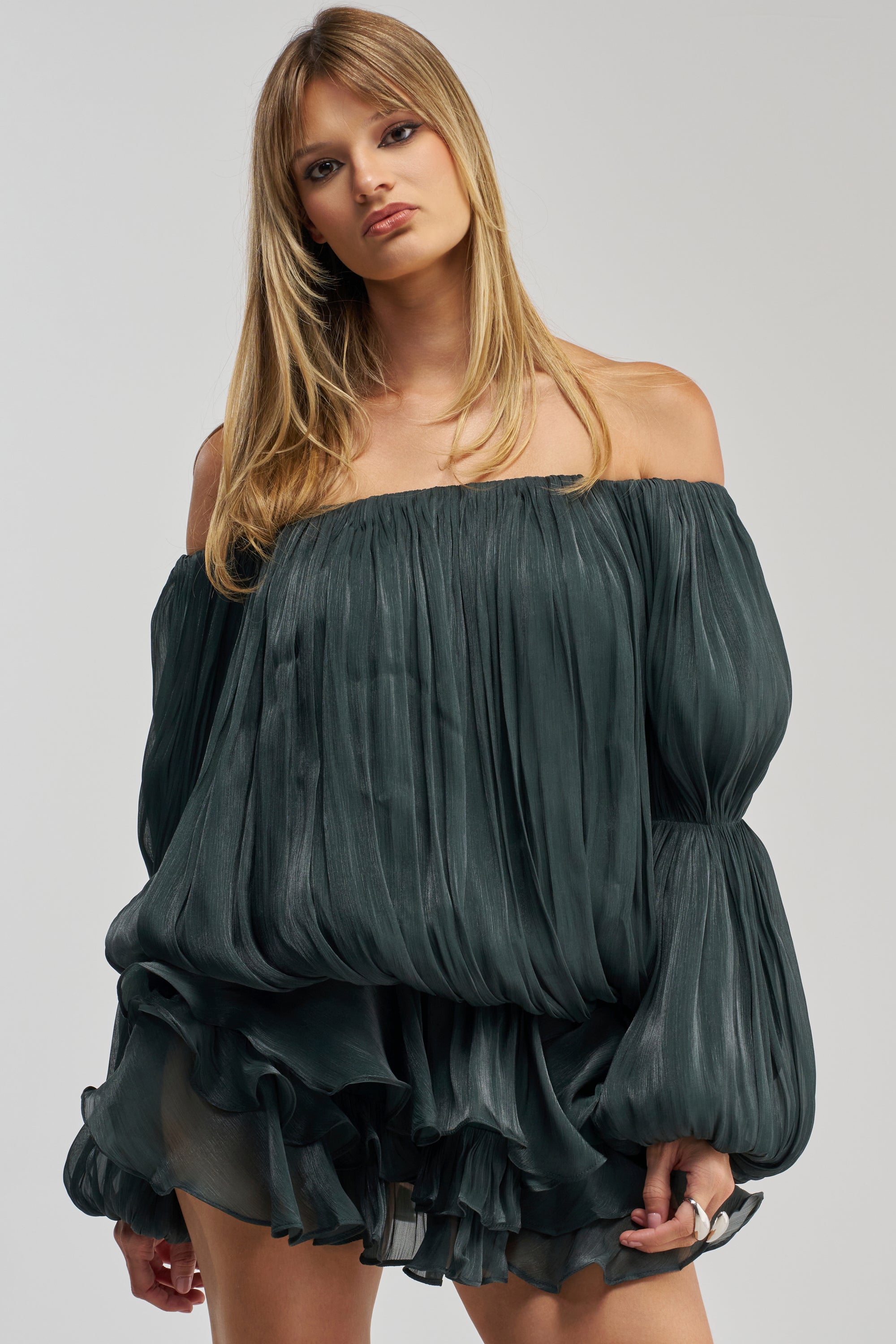 A woman with straight blonde hair wears the FAVORITE DAUGHTER Chiffon Mini Dress in Deep Sea. This off-the-shoulder dress features pleated, voluminous details and long puffed sleeves, evoking the style of a ruffle mini dress. She stands against a plain light background, gazing directly at the camera.