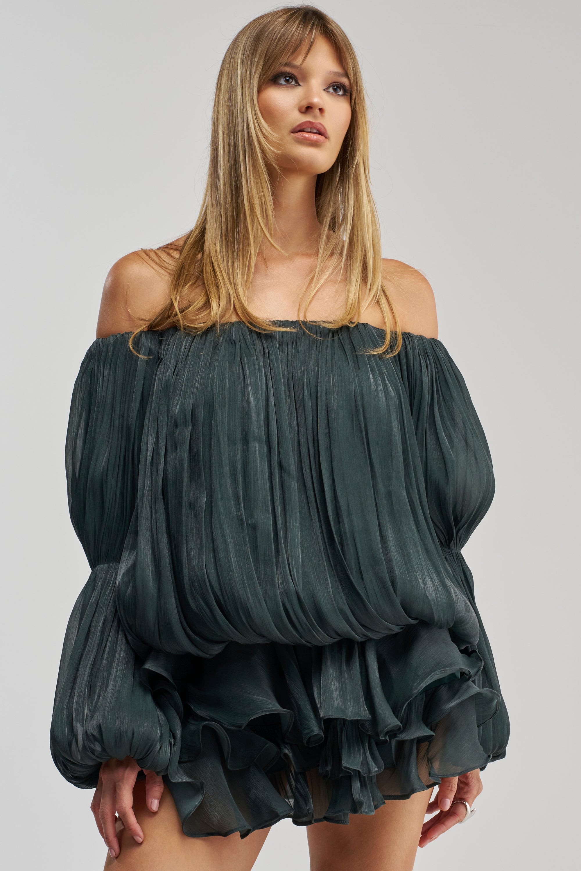 A woman stands against a plain light background, gazing slightly upward, wearing the FAVORITE DAUGHTER CHIFFON MINI DRESS IN DEEP SEA. The dress features an off-the-shoulder neckline and a voluminous pleated skirt, while its ruffled hem adds movement and texture to the elegant ensemble.