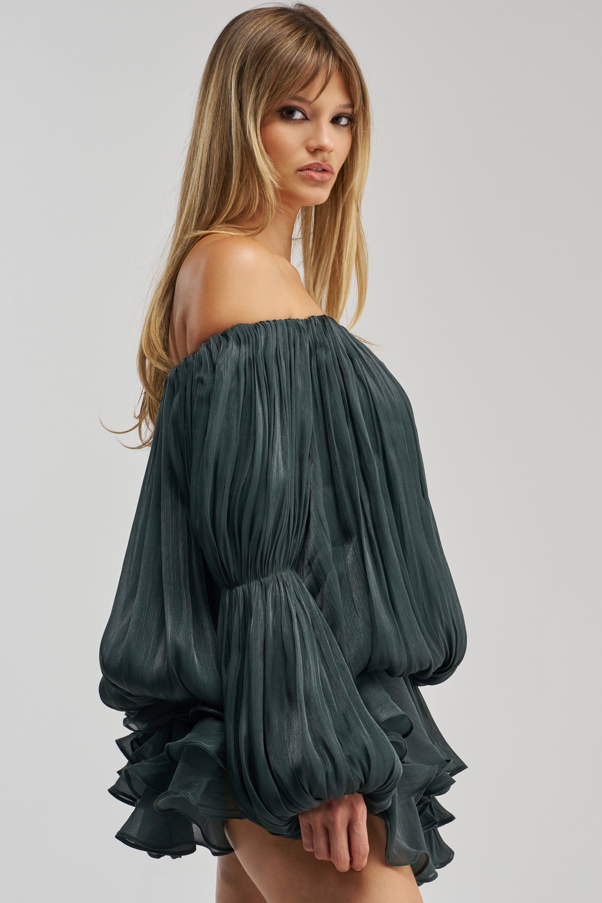 A woman with long, straight blonde hair models the FAVORITE DAUGHTER CHIFFON MINI DRESS IN DEEP SEA—a dark green, off-the-shoulder mini dress featuring voluminous pleated sleeves and a pleated skirt—as she stands against a plain light background.